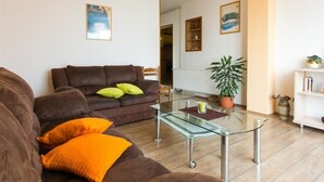 Una sala de estar con un sofá marrón, cojines naranja, una mesa de centro de vidrio y una planta en maceta.