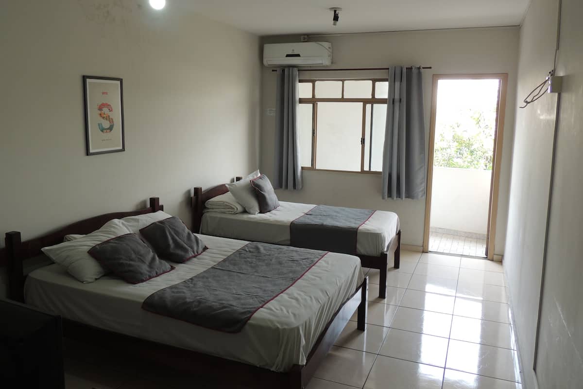 A hotel room with two beds, a ceiling fan, an air conditioning unit, a window with curtains, and a tiled floor.