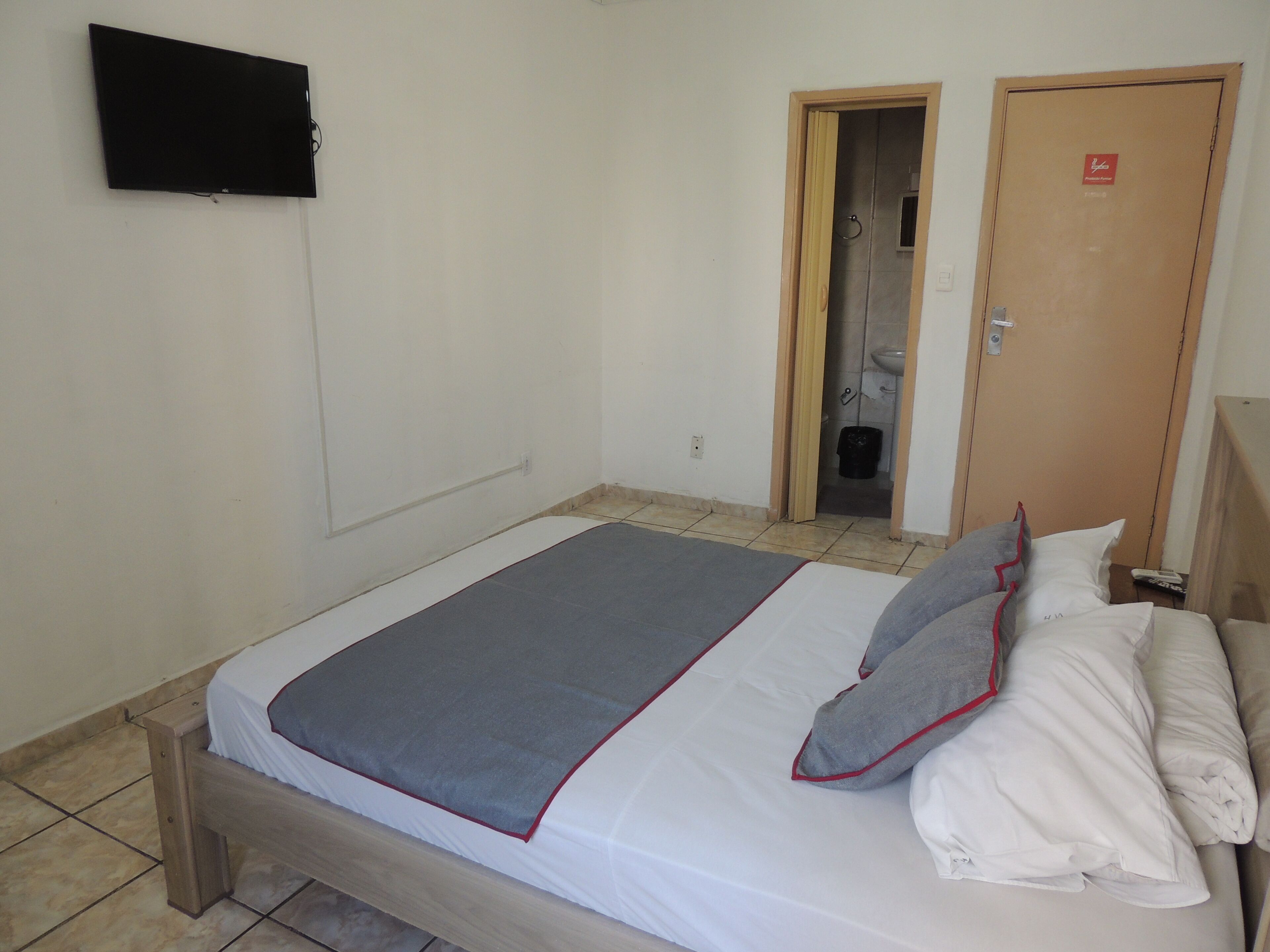 Une chambre avec un lit simple, un cadre en bois, une couverture grise, du linge de lit blanc et des oreillers gris bordés de rouge.