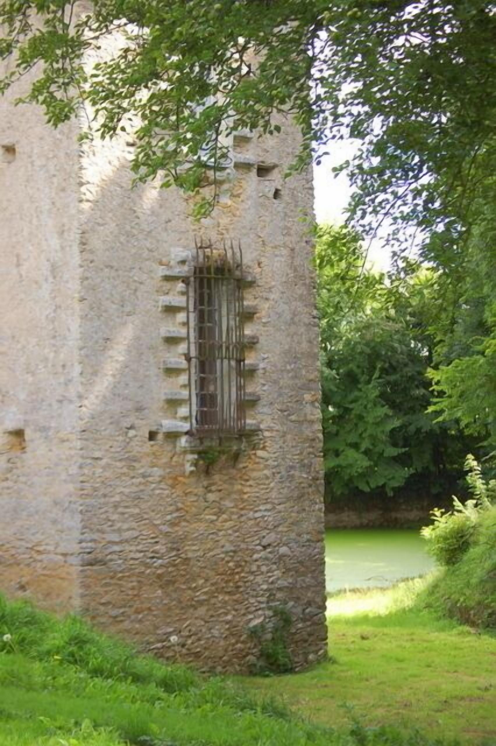 A stone tower with a barred window, surrounded by greenery and a body of water.