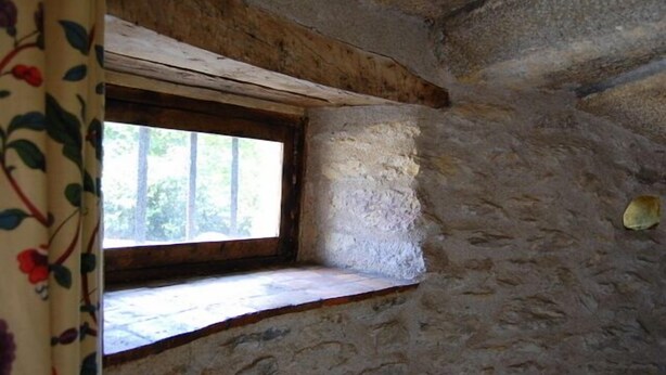 Quarto com paredes de pedra, uma pequena janela e teto com vigas de madeira.