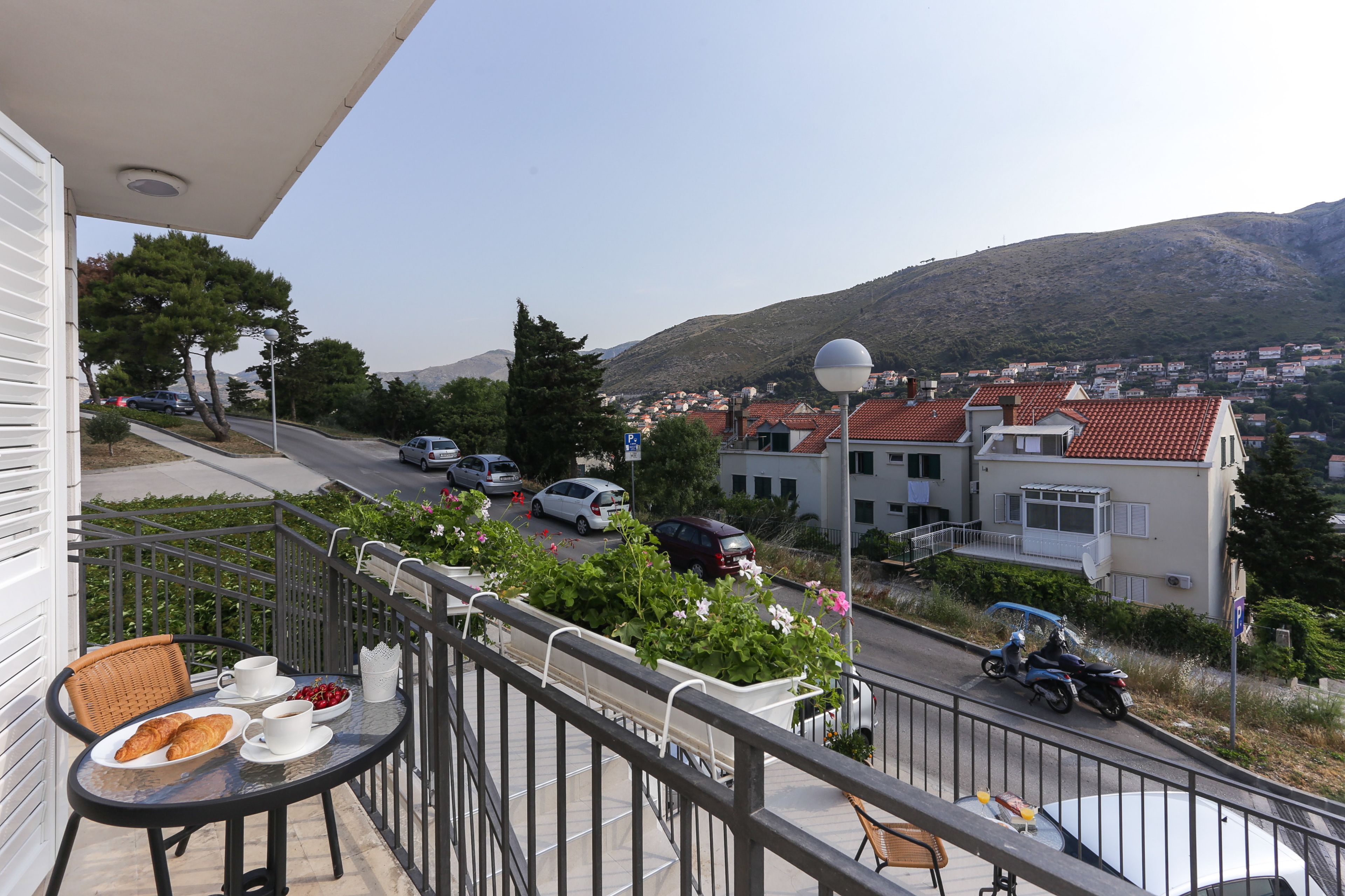Un balcon avec une table dressée pour le petit-déjeuner, donnant sur une rue animée avec des voitures et des maisons, et une montagne en arrière-plan.