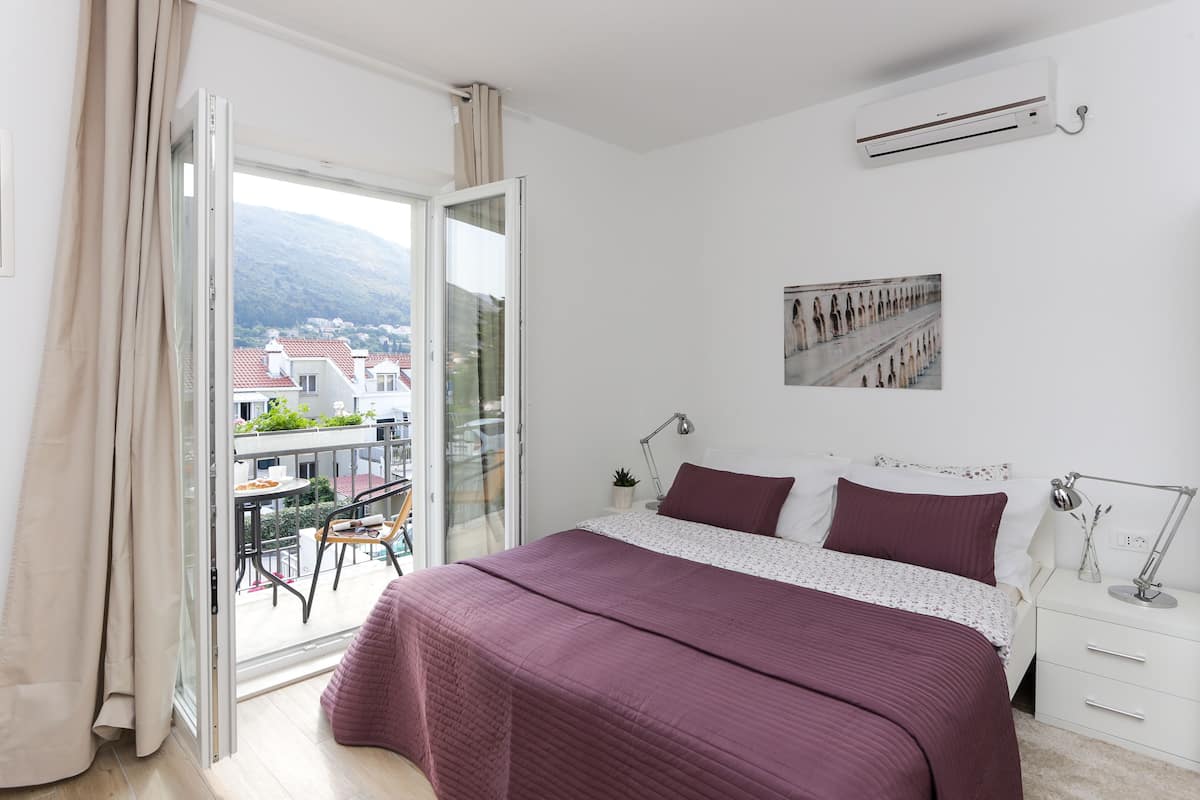 A bedroom with a large bed, a nightstand, a desk, and a chair. There is a window with curtains, a picture on the wall, and an air conditioning unit.