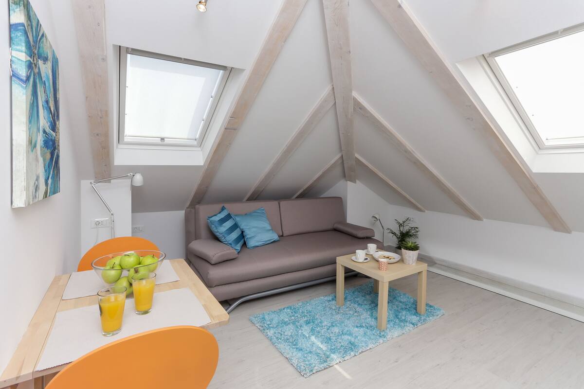 A modern loft bedroom with a sloped ceiling, a sofa, a dining table with fruit and drinks, and a small side table with a plant.
