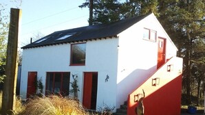 A white building with red accents, including doors and a staircase, surrounded by natural vegetation. - The Village Studio Apartments (Moate)