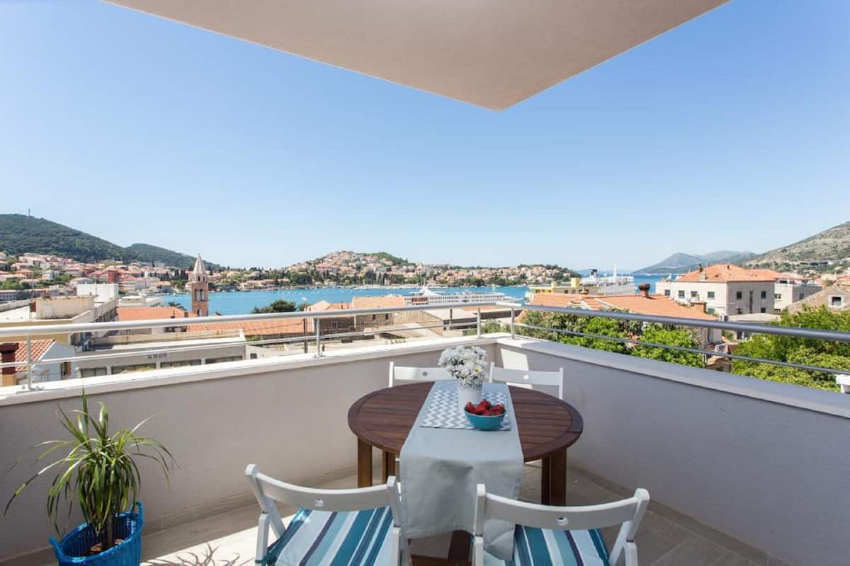 Varanda com mesa e cadeiras, vista para uma vila costeira, com mar e colinas visíveis.