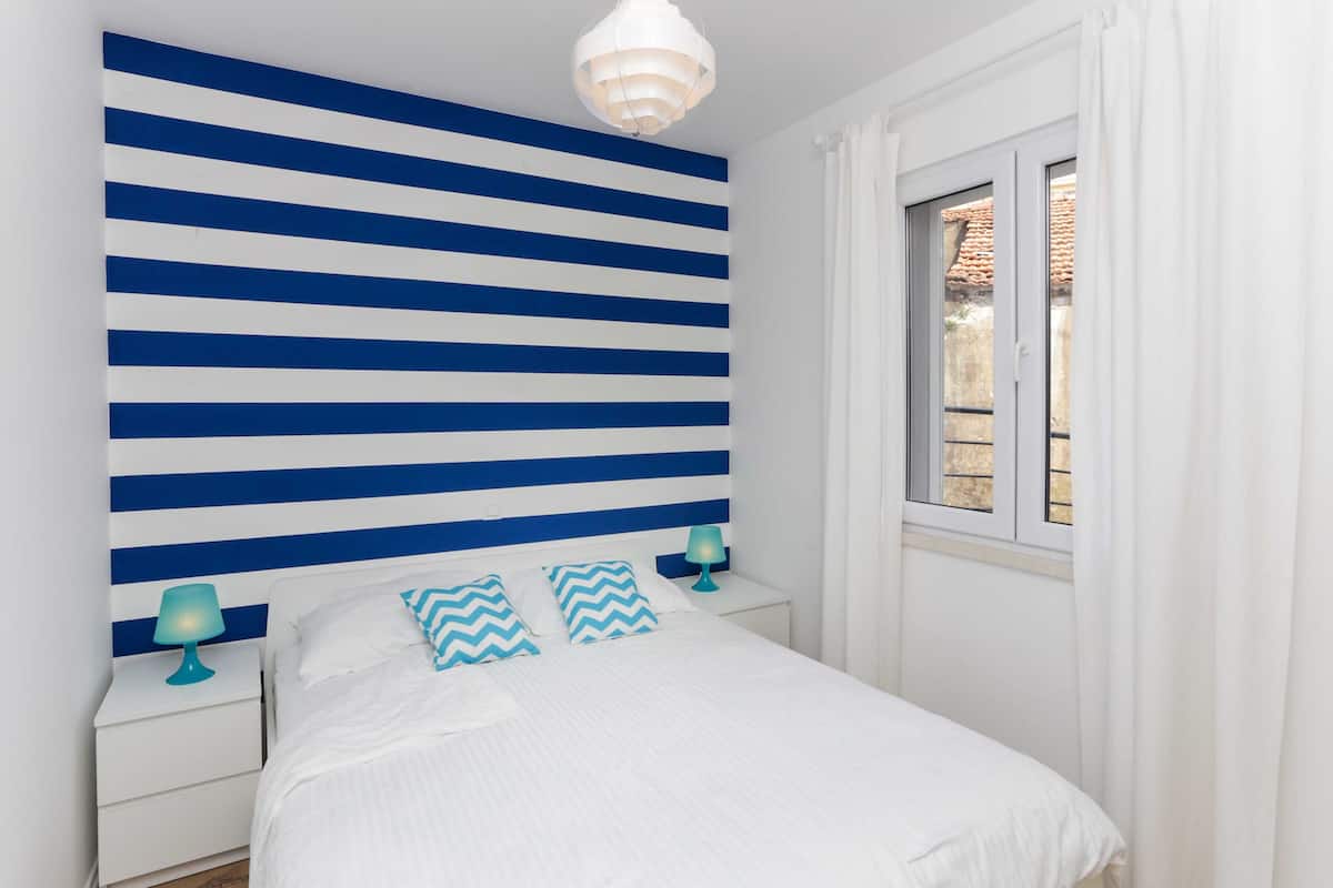 Quarto com cabeceira azul e branca listrada, cama branca com almofadas combinando, mesa de cabeceira com abajur e janela com cortinas.