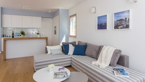 A modern living room with a grey and white striped sofa, a round coffee table with magazines, and a kitchen area in the background. - Apartments Villa Karmen (Dubrovnik)