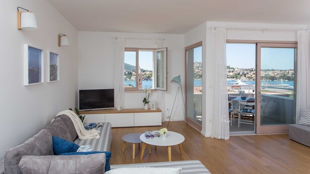 A modern living room with a sofa, a TV, a small table, and a balcony with a view of the city and water. - Apartments Villa Karmen (Dubrovnik)