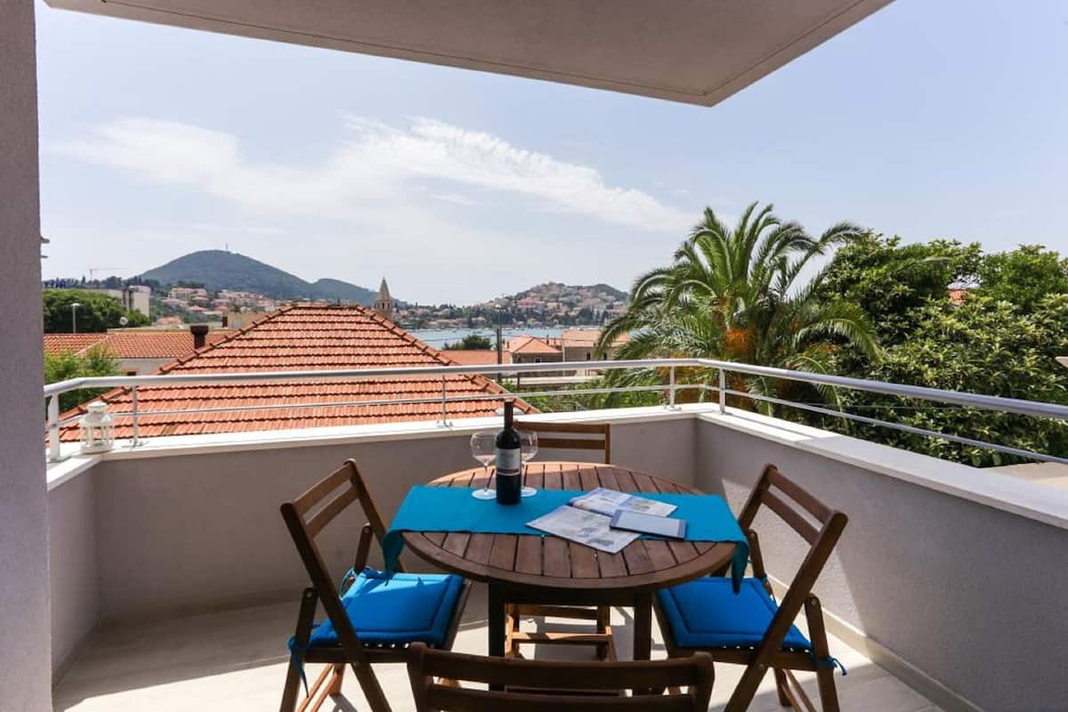 Uma varanda com uma mesa de madeira redonda, duas cadeiras com almofadas azuis e uma garrafa de vinho.