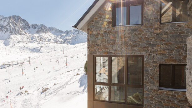 A stone building with large windows, a ski slope in the background, and a clear sky.