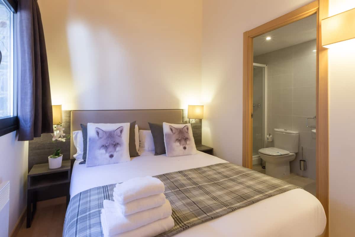 Une chambre d’hôtel avec un lit soigneusement fait, une salle de bain visible à travers une porte ouverte, et une table de chevet avec une lampe et une petite plante en pot.
