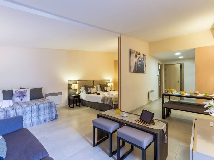 A modern hotel room with a bed, a sofa, a desk with a laptop, and a dining area with a table and chairs.