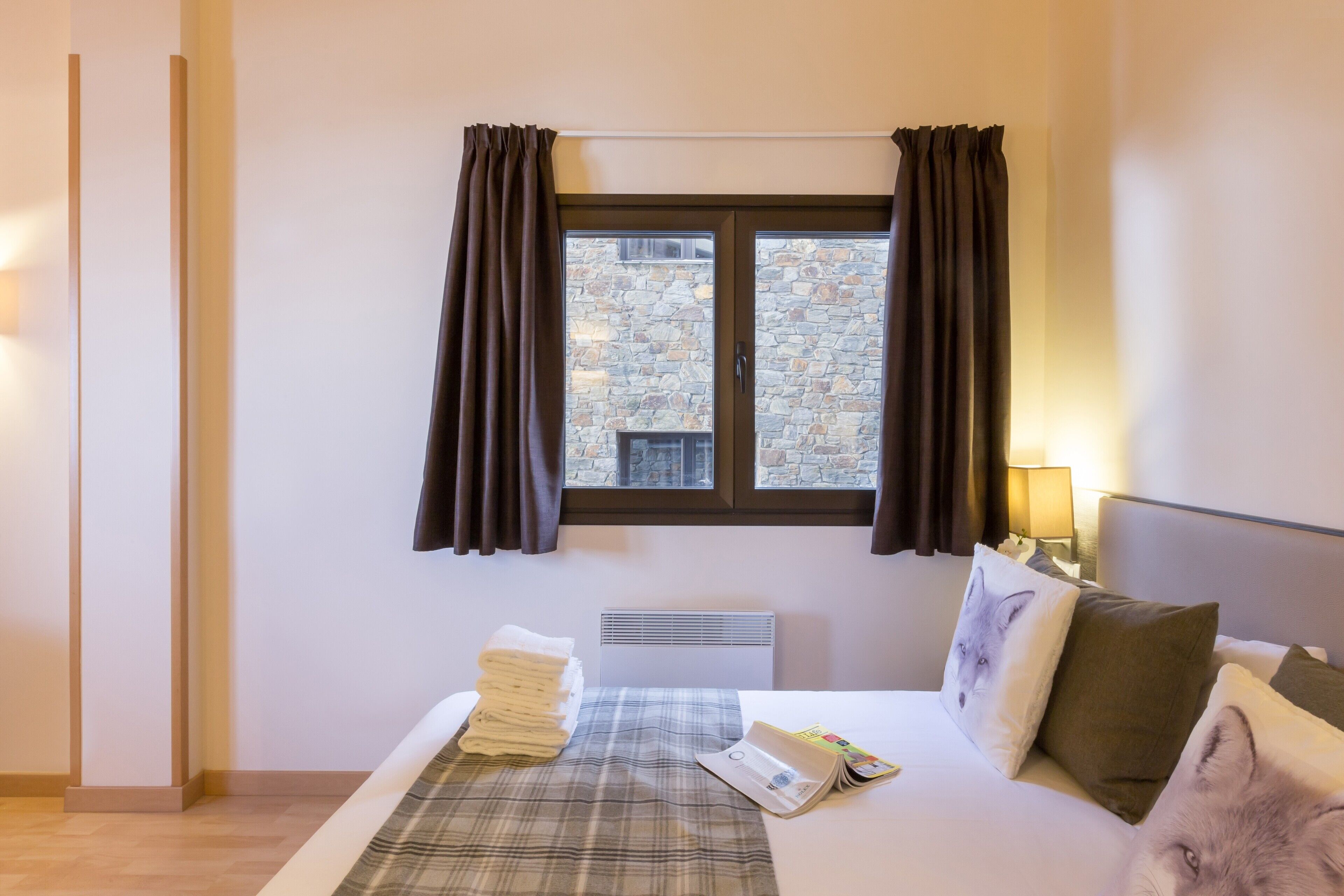 A hotel room with a bed, a window with curtains, a radiator, and a stack of towels on the bed.