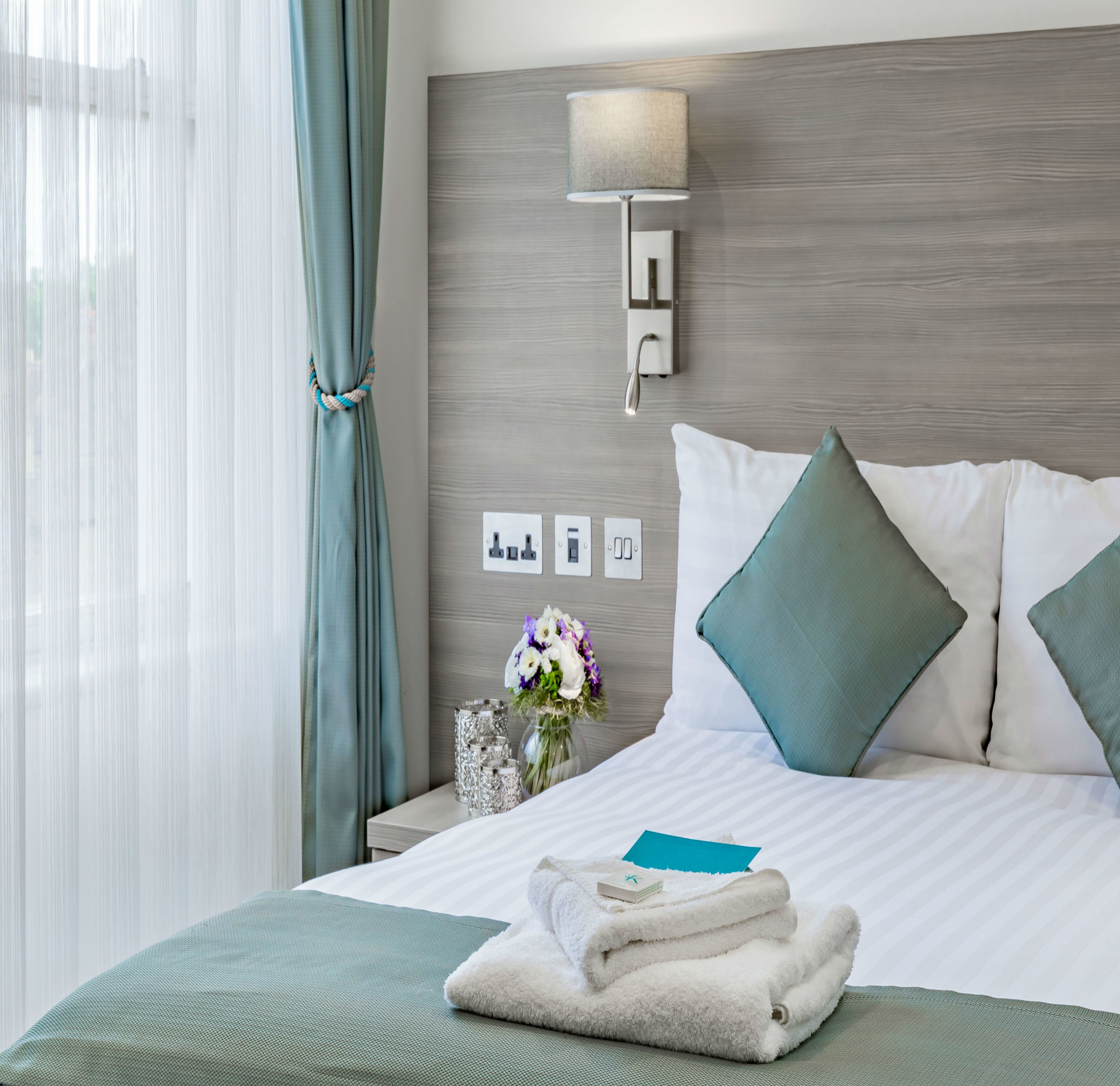 A neatly made bed with a folded towel, a bedside lamp, and a vase with flowers.