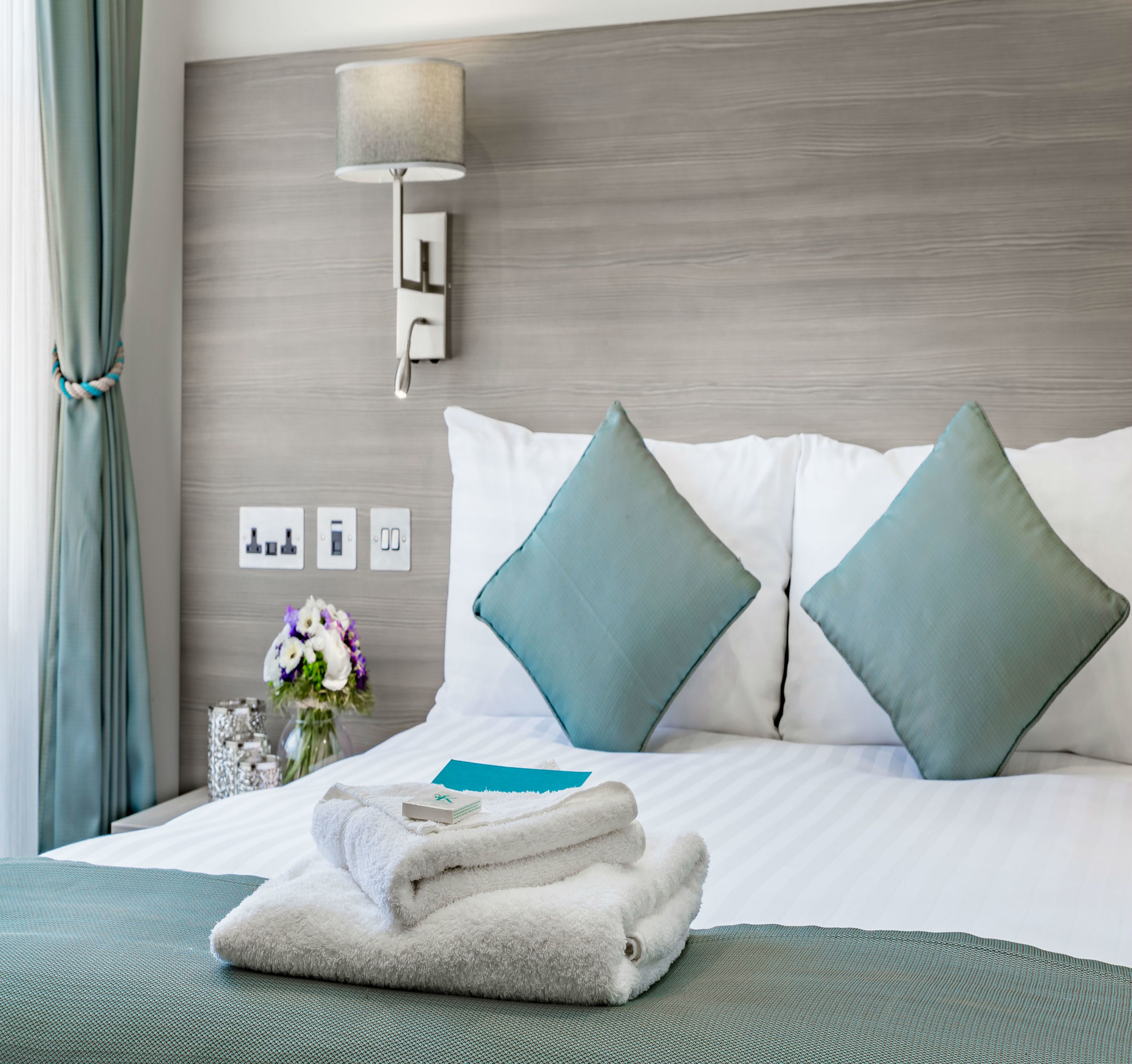 A neatly made bed with two pillows, a bedside lamp, a vase with flowers, and a set of folded towels.