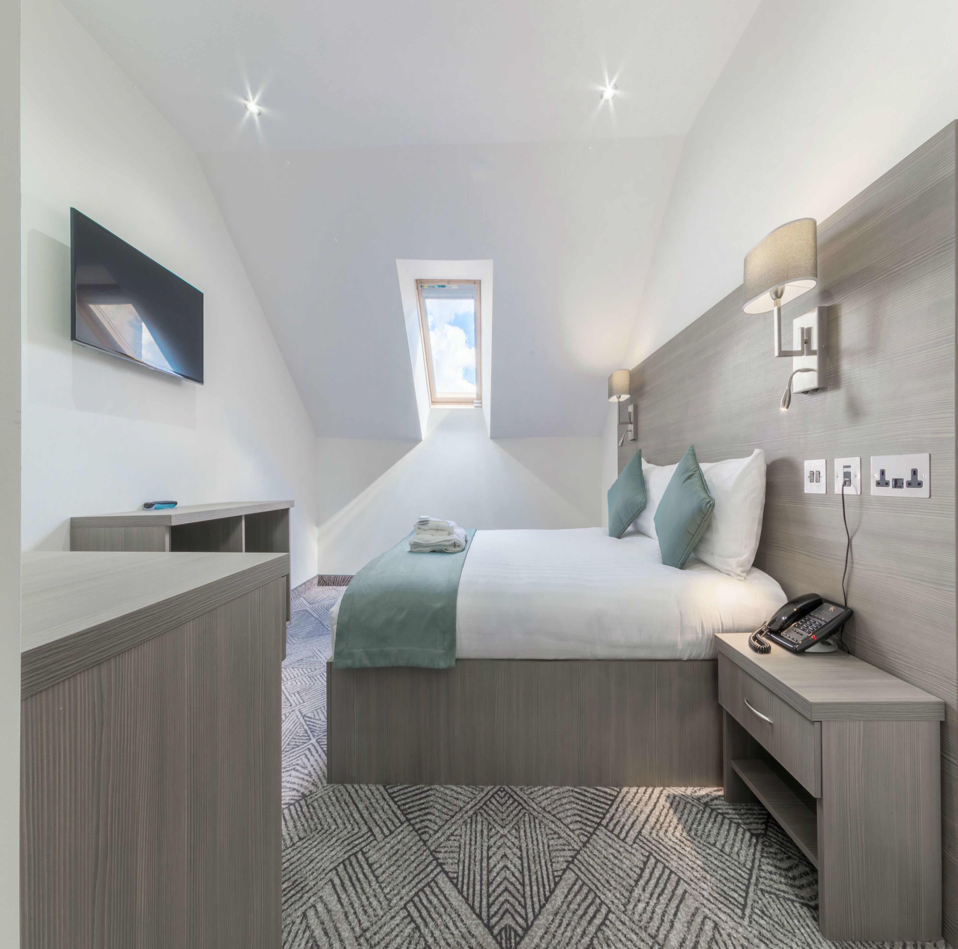 Un dormitorio moderno con cama, mesita de noche, lámpara y televisor montado.
