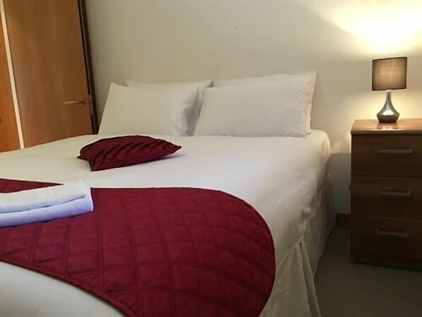 Cama bem arrumada com manta vermelha e almofadas, luminária de cabeceira e mesa de cabeceira de madeira com gavetas.