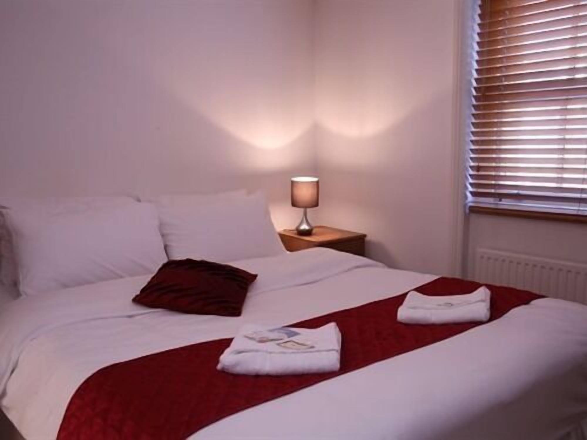 A neatly made bed with white and red bedding, a bedside lamp, and a window with blinds.