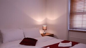 Una cama bien hecha con ropa de cama blanca y roja, una lámpara de noche y una ventana con persianas.