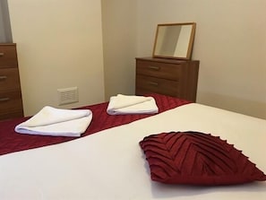 Quarto de hotel com cama, duas toalhas brancas dobradas, um travesseiro vermelho e cômoda de madeira com espelho.