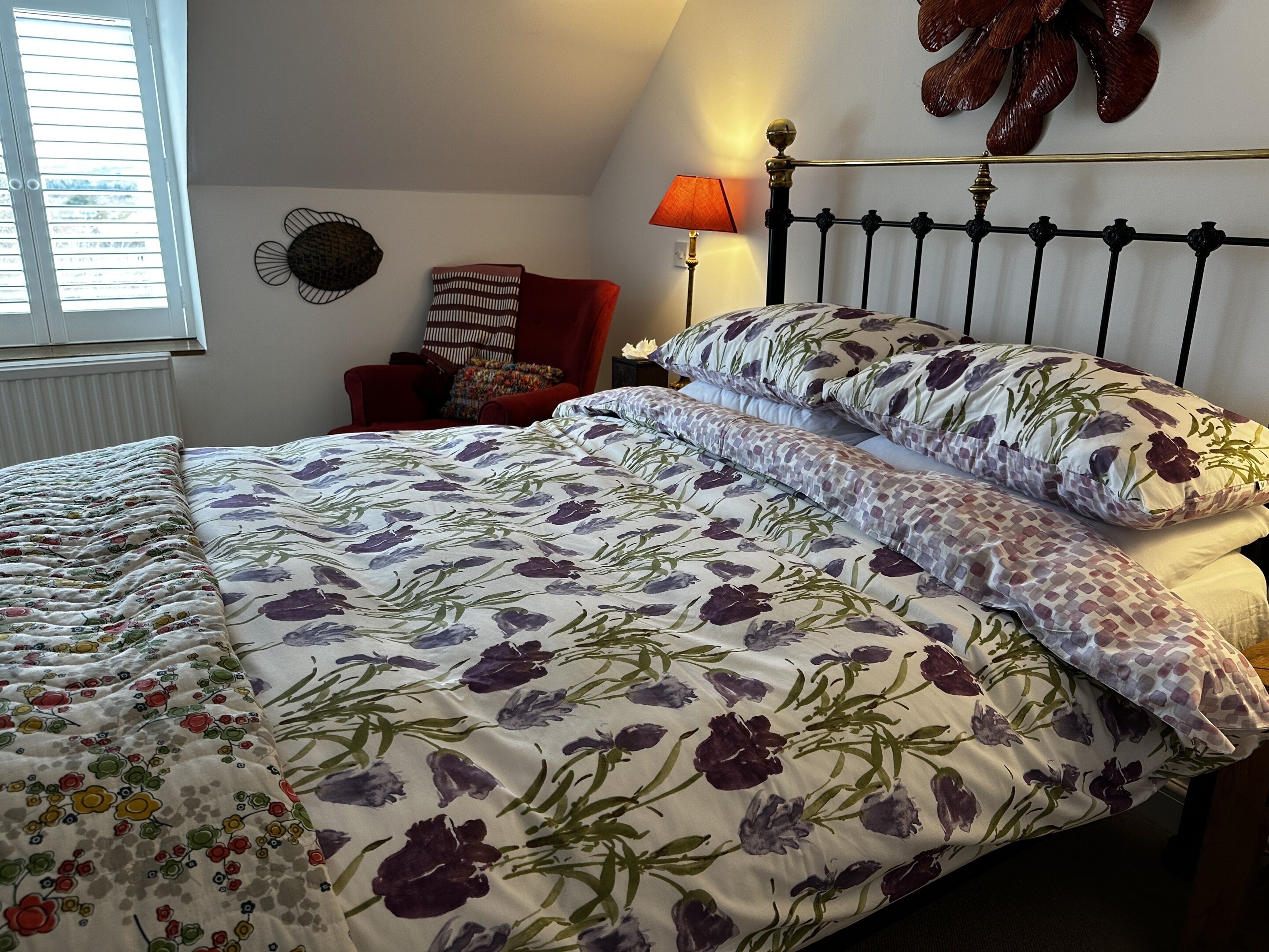 A neatly made bed with floral bedding, a bedside table with a lamp, and a chair with a patterned cushion.