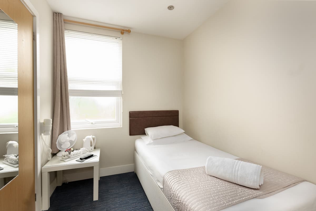 A small, neatly arranged hotel room with a single bed, a bedside table with a lamp, a mirror, and a window with curtains.