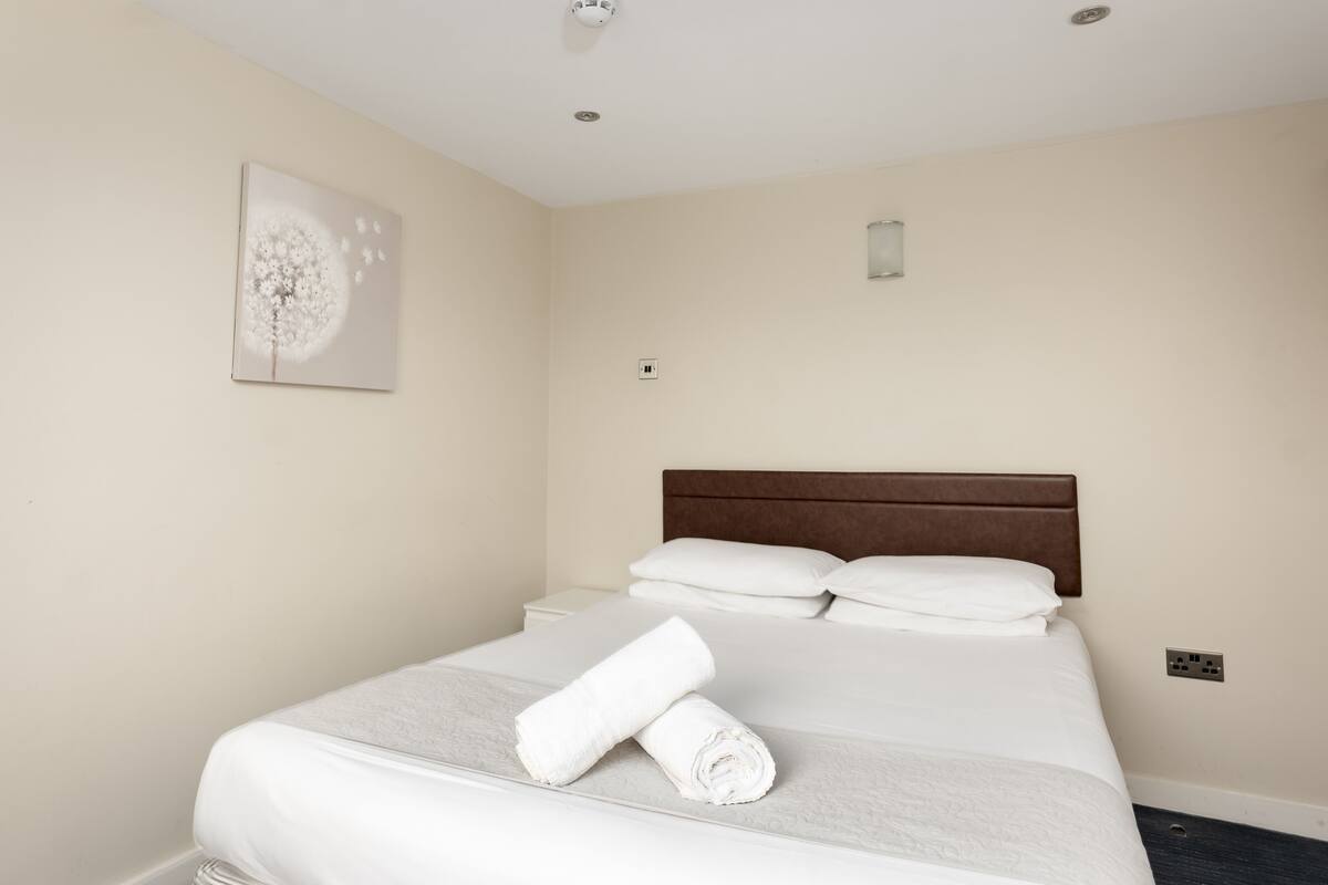 A neatly made bed with white linens and a rolled towel, a wooden headboard, and a single wall-mounted light fixture.