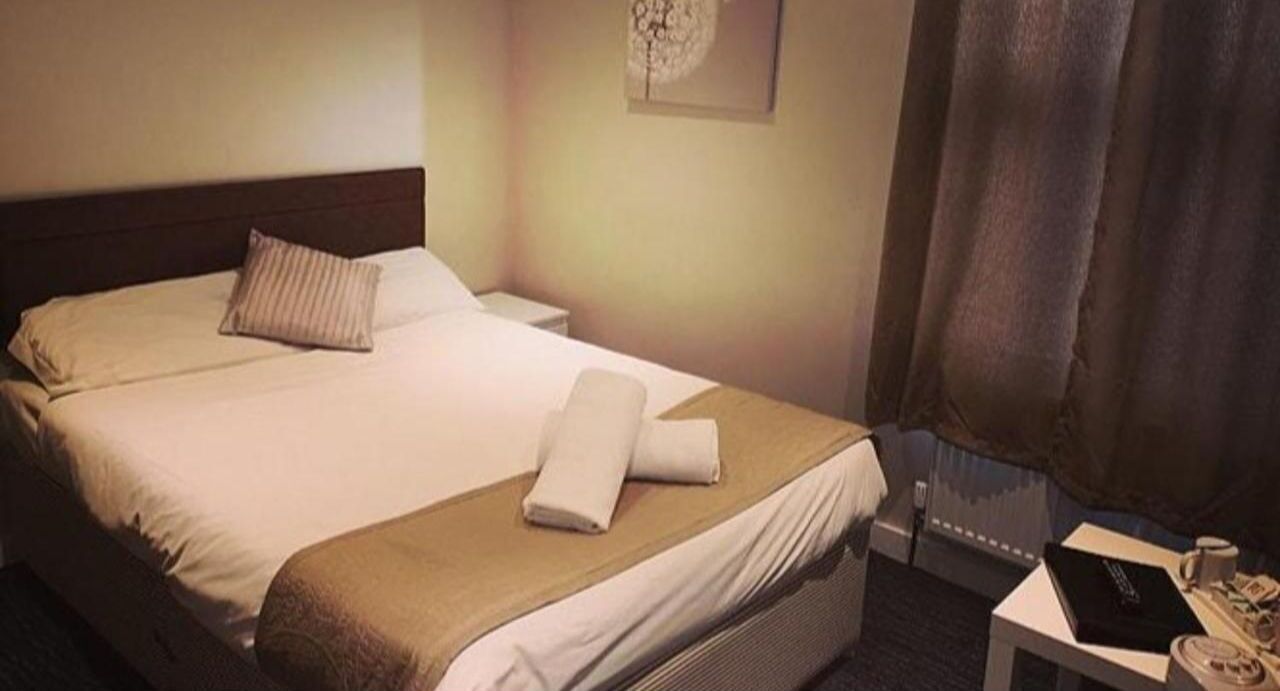 A hotel room with a bed, a bedside table with a phone and a book, and a window with curtains.
