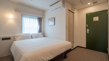 A hotel room with a bed, a window with curtains, an air conditioning unit, a green door, and a wall-mounted light.