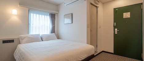 A hotel room with a bed, a window with curtains, an air conditioning unit, a green door, and a wall-mounted light.