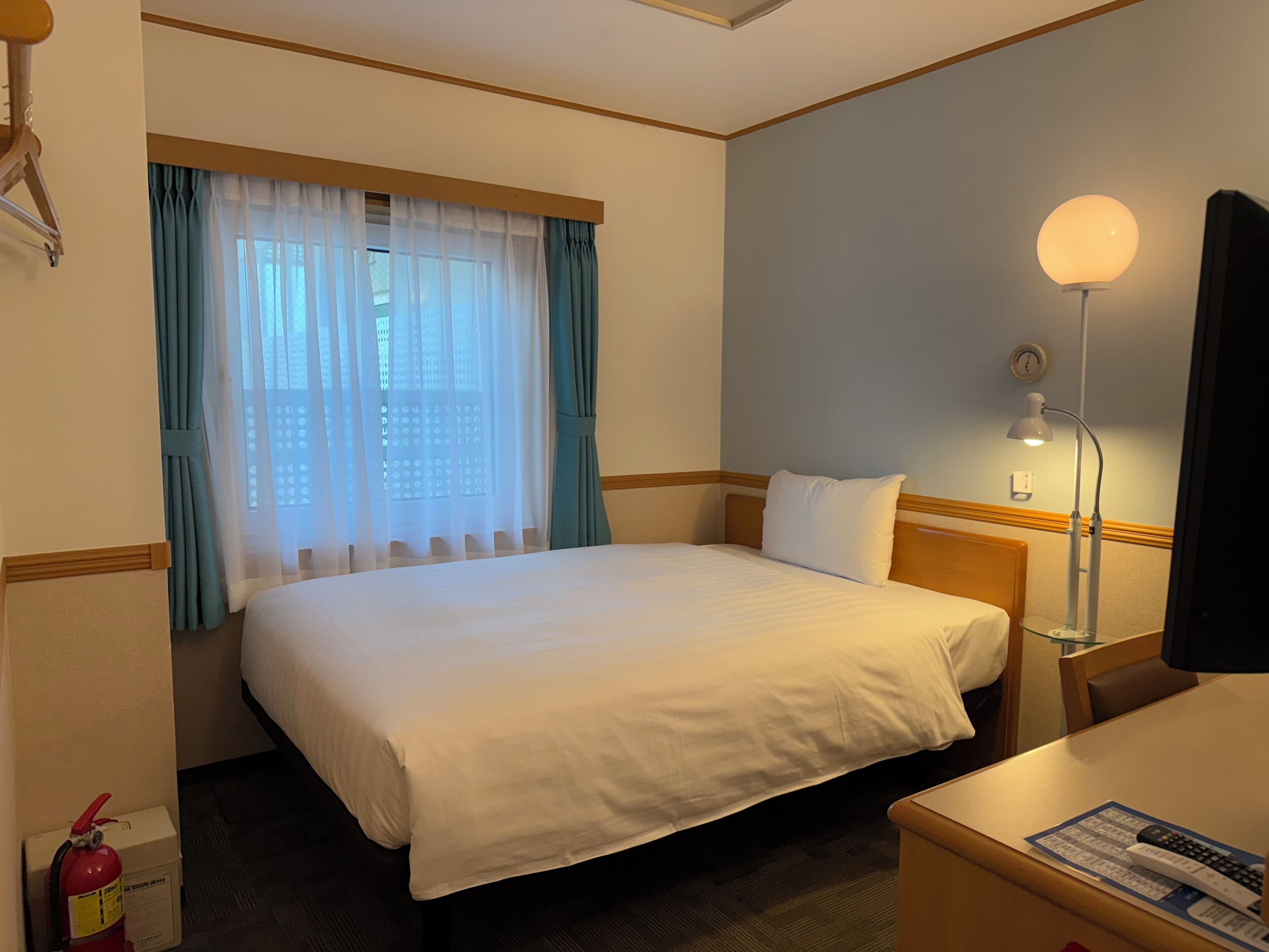 A hotel room with a bed, a desk, a lamp, a fire extinguisher, and a window with curtains.