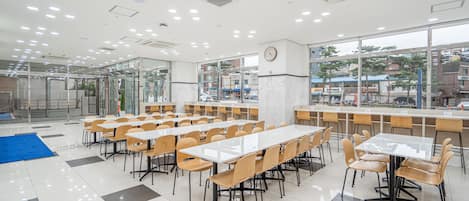 Una amplia cafetería con numerosas mesas y sillas, grandes ventanales que ofrecen vistas de la ciudad, y un reloj en la pared.