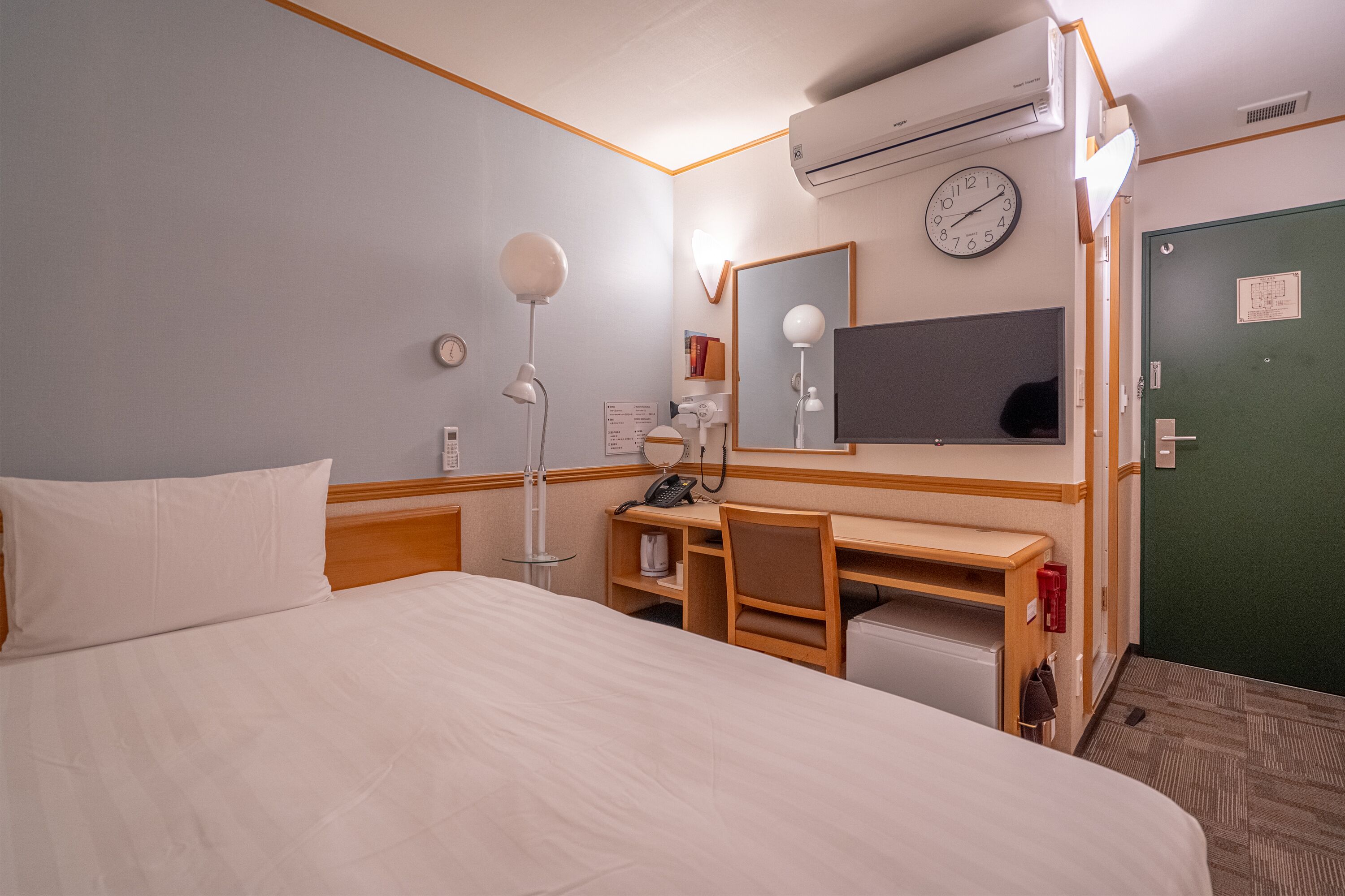 Et hotelværelse med en stor seng, et skrivebord med stol, en skrivebordslampe, en telefon, et spejl og en grøn dør.