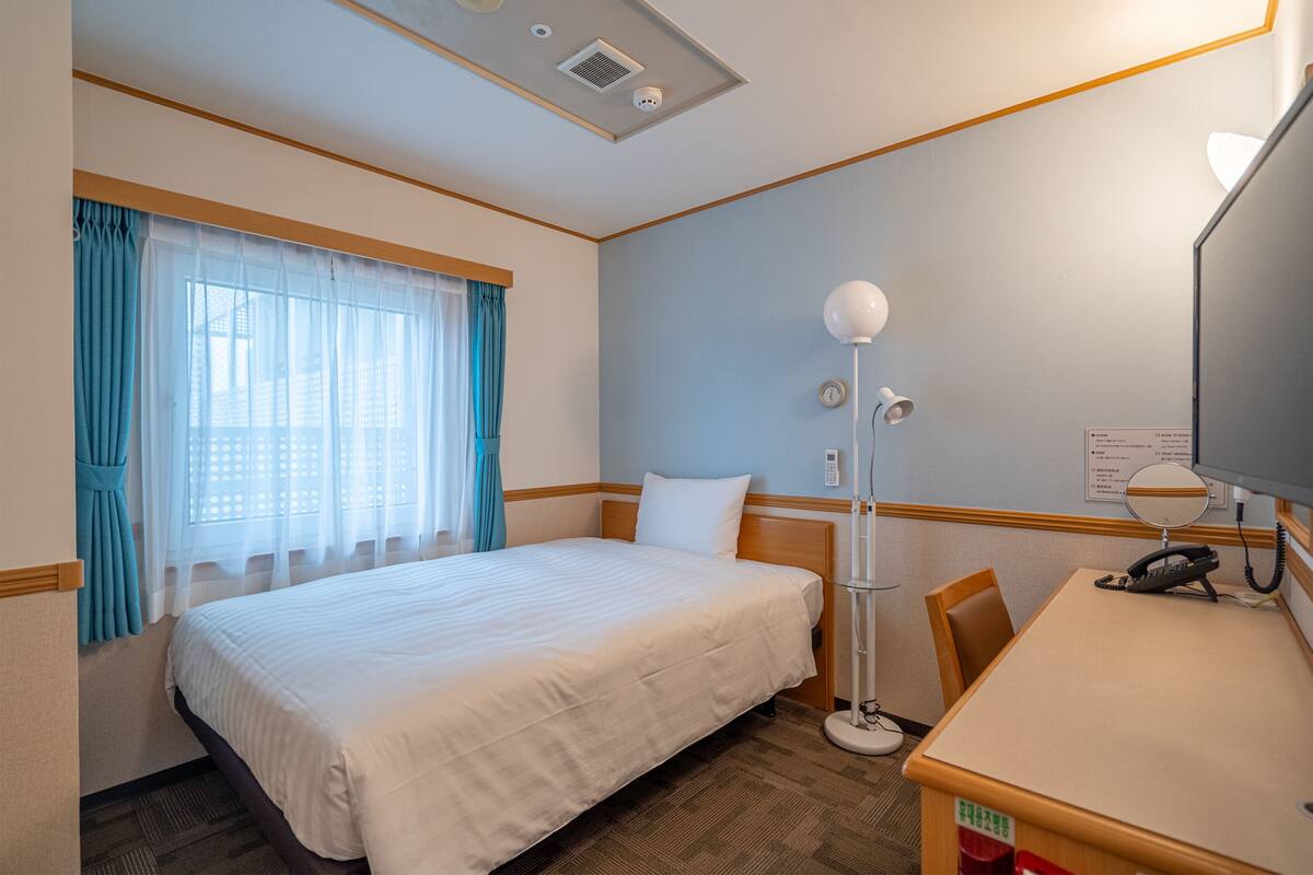 Une chambre d’hôtel avec un lit, un bureau avec un téléphone, une lampe et une fenêtre avec des rideaux.