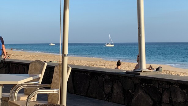 A beachside cafe with outdoor seating, a stone wall, and a view of the sea with boats. - Apartamentos Alberto (Pajara)