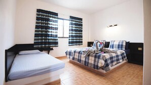 A room with two beds, a window with striped curtains, a bedside table, and a wall-mounted light fixture. - Apartamentos Alberto (Pajara)