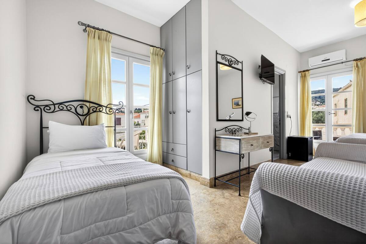 Quarto com duas camas, guarda-roupa, espelho, escrivaninha e janela com cortinas.