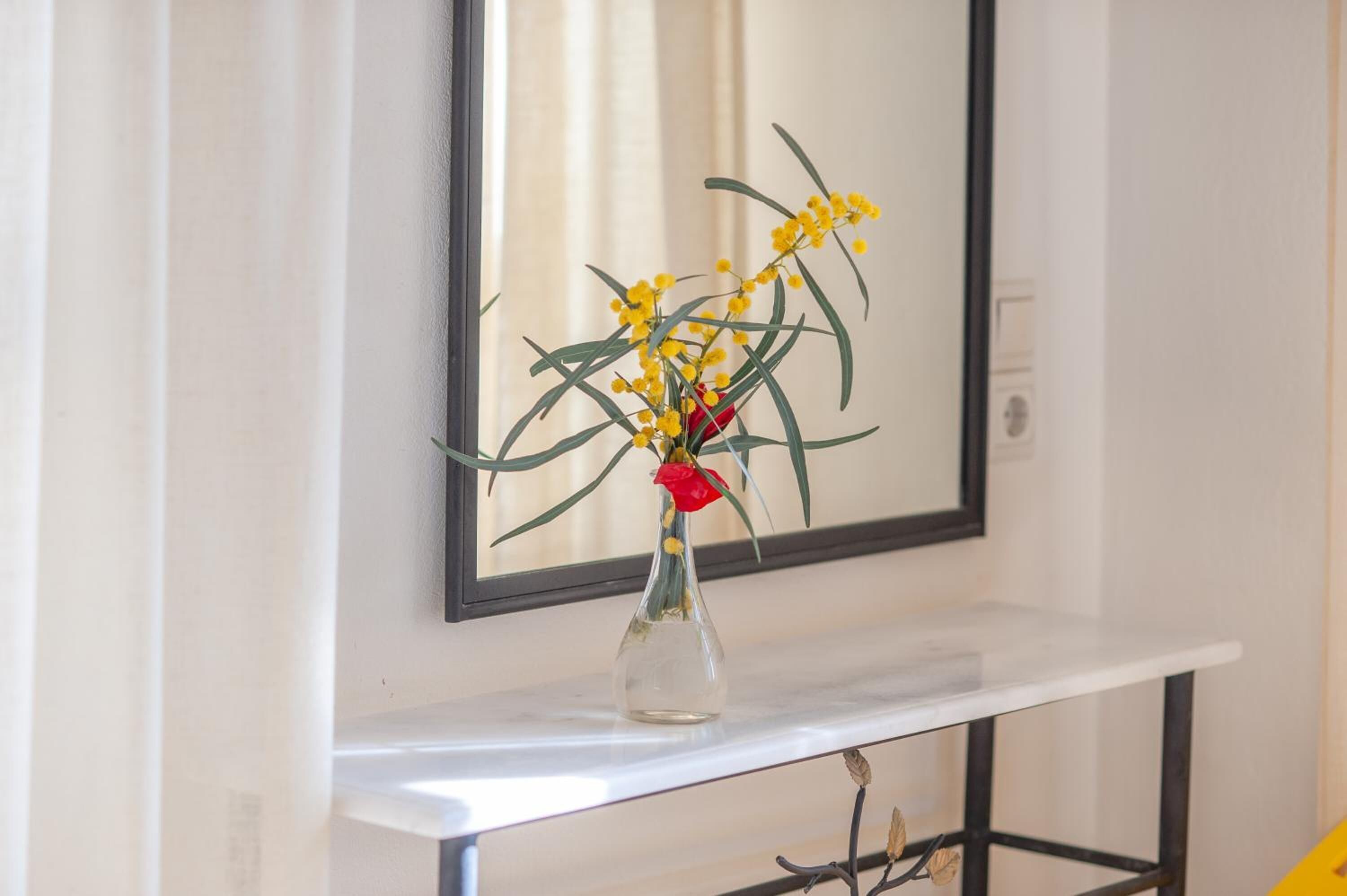 A clear view of a vase with yellow flowers and a red rose on a white shelf against a wall with a mirror.