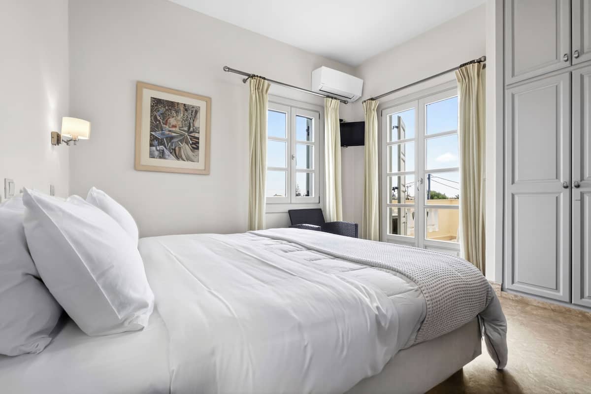 Quarto com cama, uma cadeira, um quadro e vista pela janela.
