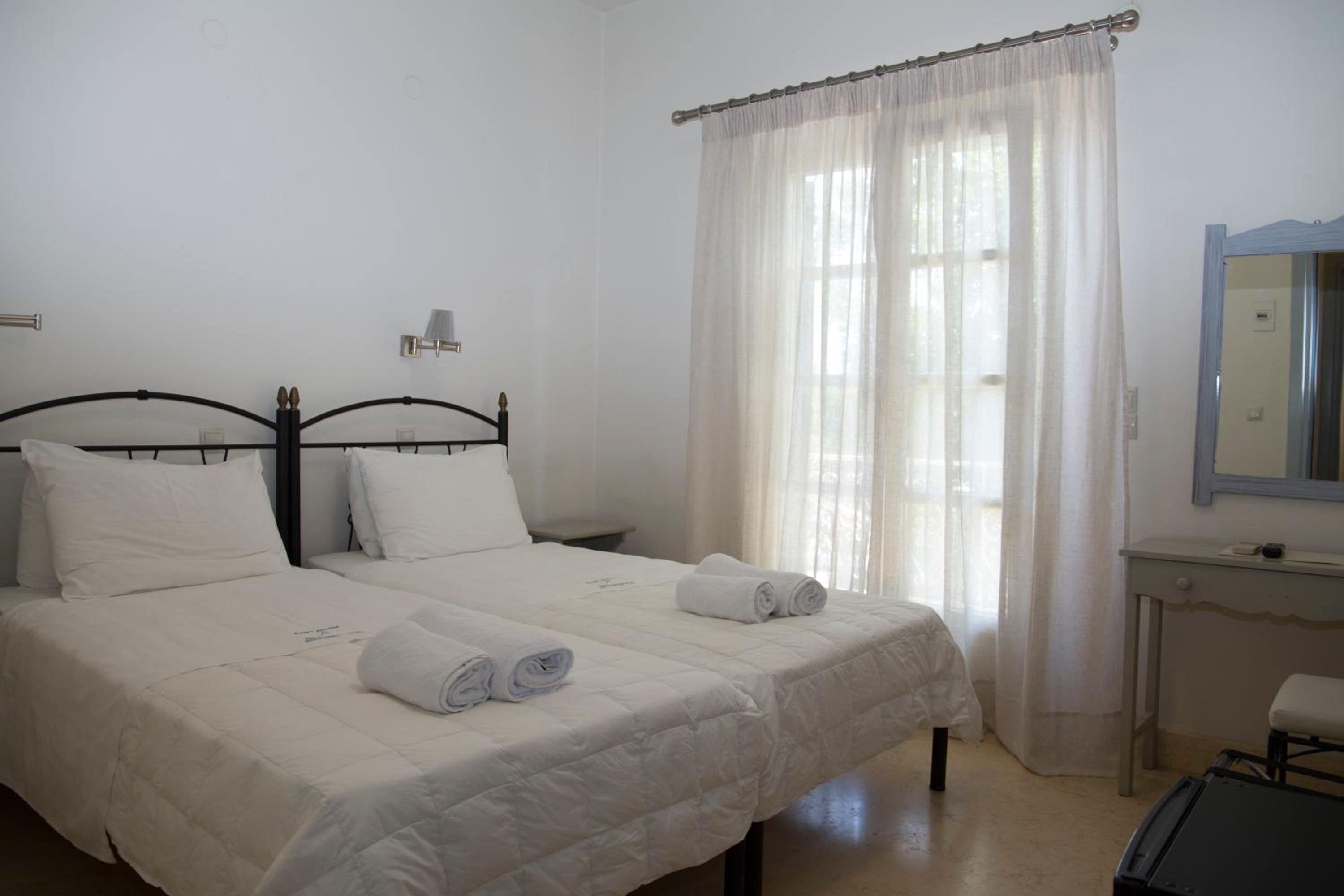 Habitación de hotel con dos camas, una mesita de noche, un espejo y una ventana con cortinas de gasa.