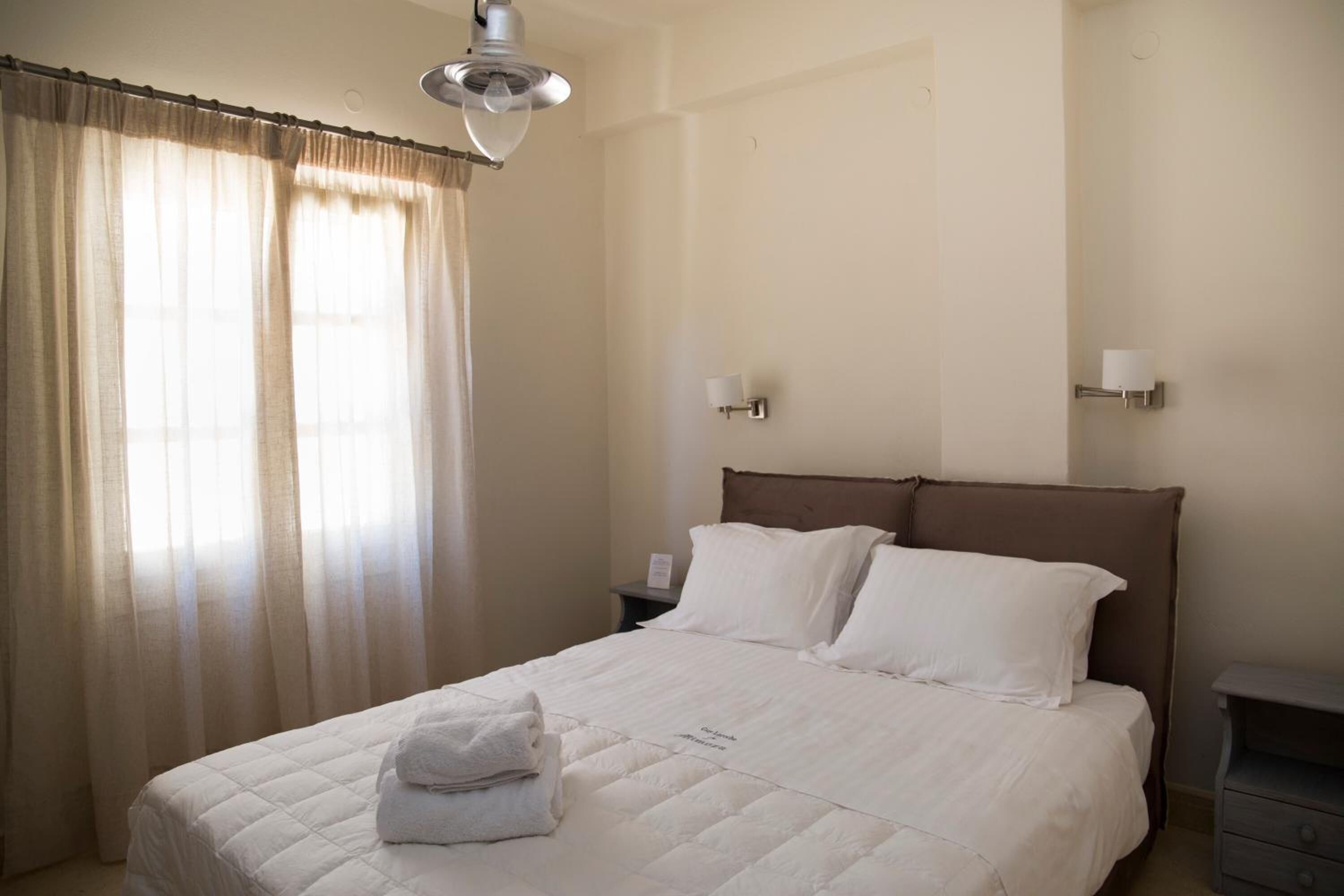 A neatly made bed with a white duvet and pillows, a bedside table with a lamp, and a window with sheer curtains.