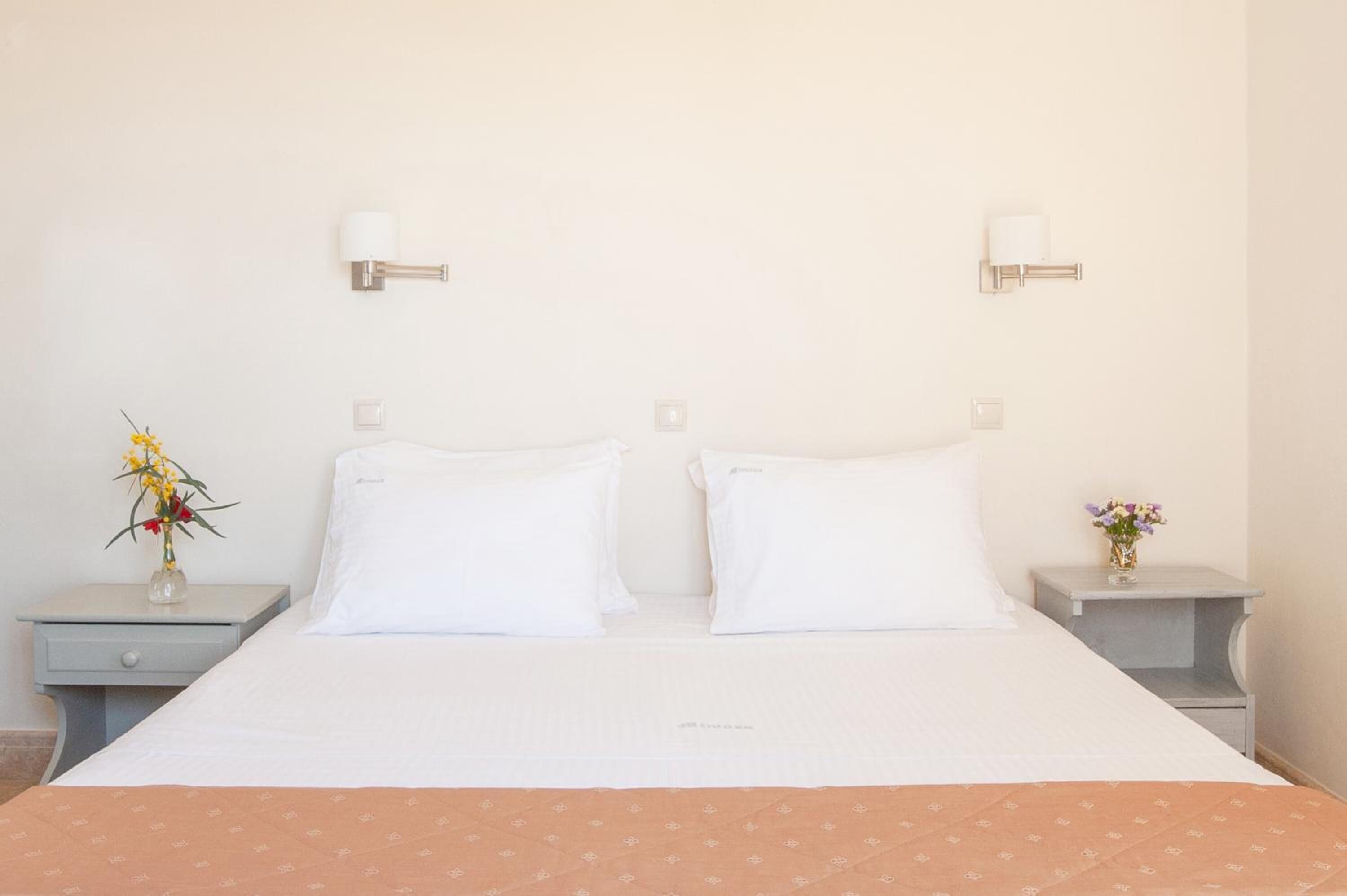 Een netjes opgemaakt bed met witte lakens en een bruin sprei, omlijst door twee nachtkastjes met lampen en bloevasen.