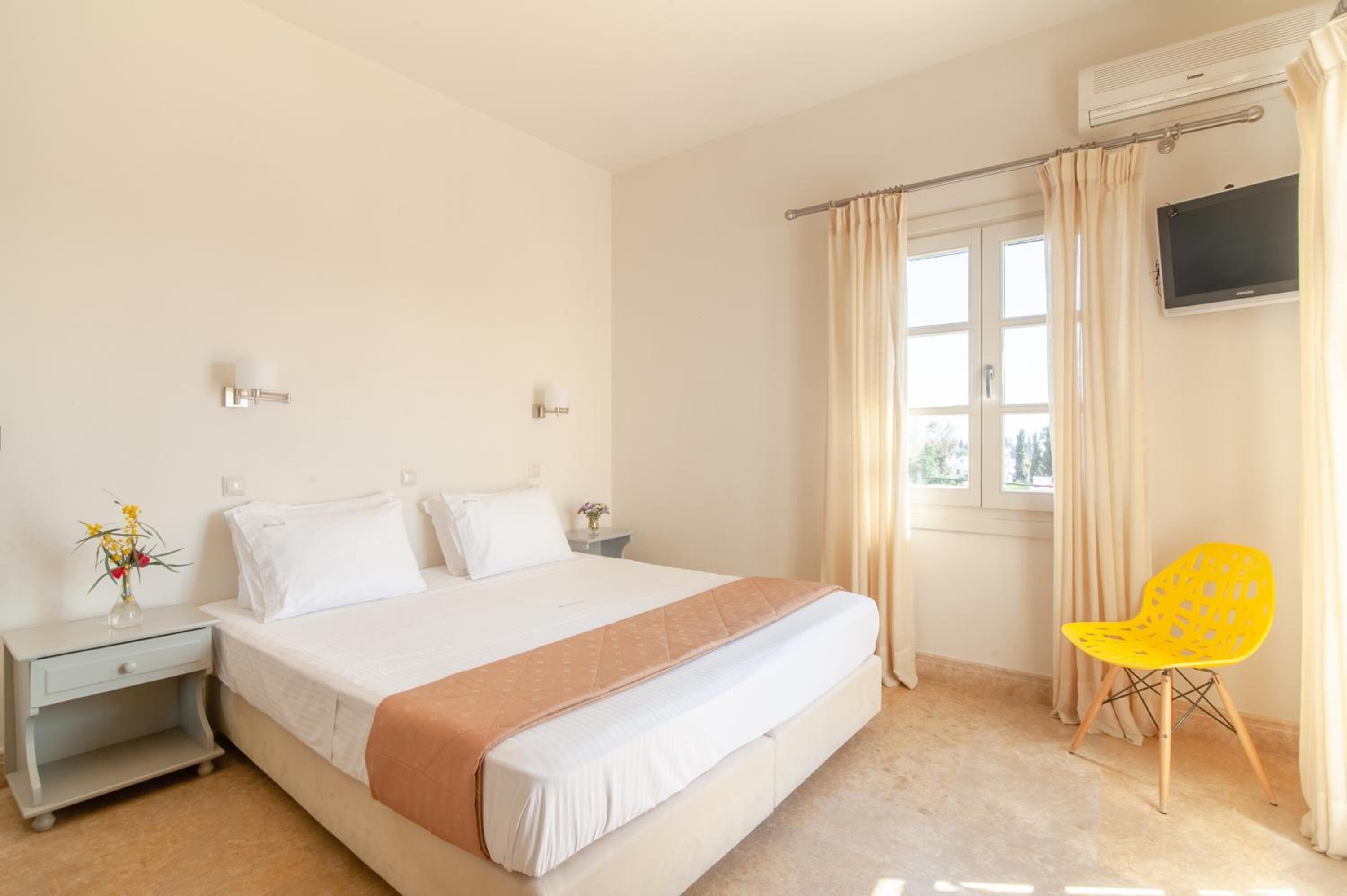 A hotel room with a large bed, a nightstand with a lamp, a TV mounted on the wall, and a window with curtains.