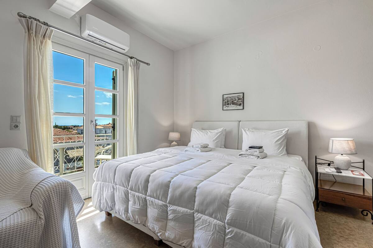 Quarto de hotel com cama, poltrona, mesa de cabeceira, luminária e janela com cortinas.