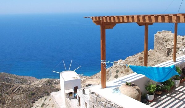 Una terraza frente al mar con una pérgola de madera, un edificio blanco y plantas en macetas.