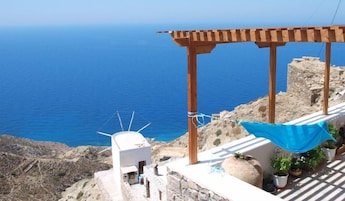 Una terraza frente al mar con una pérgola de madera, un edificio blanco y plantas en macetas.