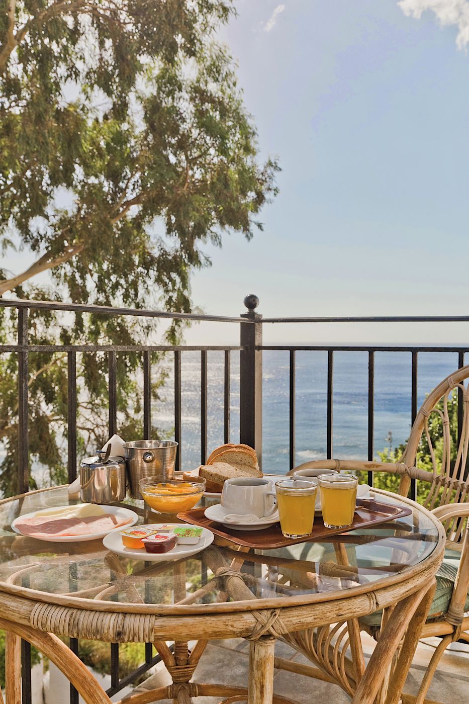 Una mesa de vidrio con alimentos para el desayuno, una silla de mimbre y vista al mar.