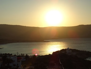 Vista del atardecer en la costa con mar tranquilo, silueta de una colina y un pequeño edificio en primer plano.
