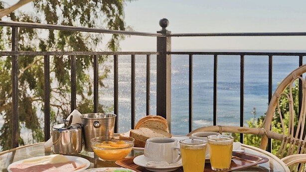 Una mesa de vidrio con alimentos para el desayuno, una silla de mimbre y vista al mar.